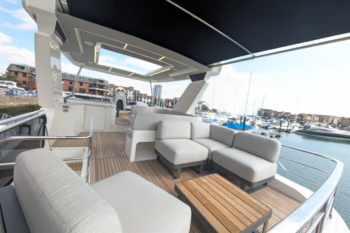 Elegant yacht deck featuring grey sofas, wooden table and marina view with moored boats and buildings in the background.